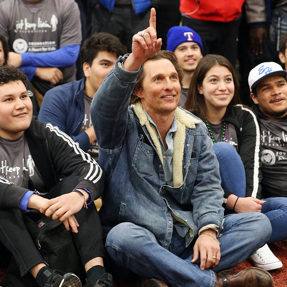Matthew Mcconaughey sitting on the floor with a group of kids holding his hand up